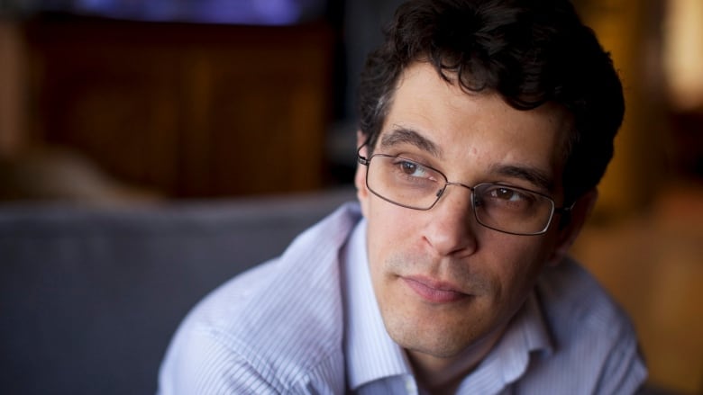 A man in spectacles and a light blue, striped shirt is seen in close-up, looking off to the left of the frame.