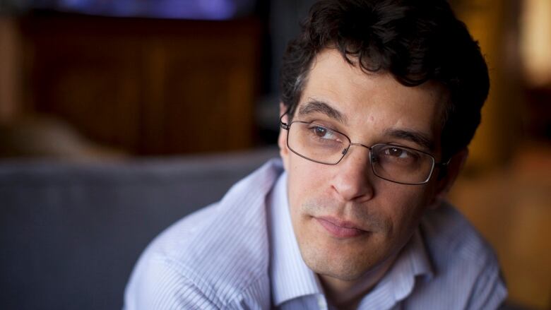 A man in spectacles and a light blue, striped shirt is seen in closeup, looking off to the left of the frame.