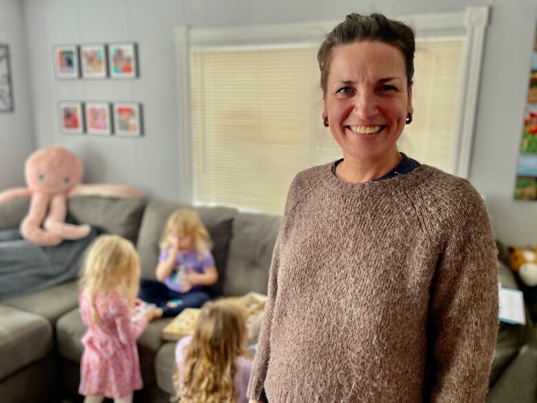 A woman with brown hair in a pony tail smiles and the camera while small children play behind her. 