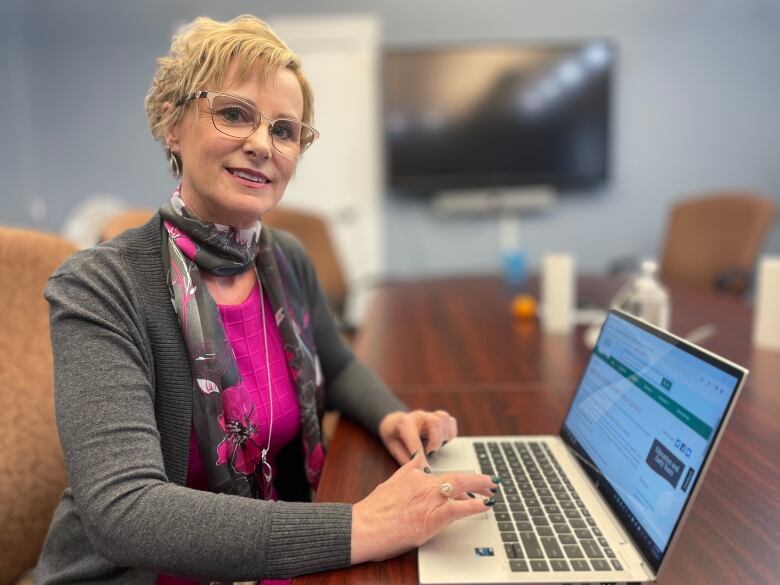 A woman with blond hair and glasses sits at a computer and smiles toward the camera.