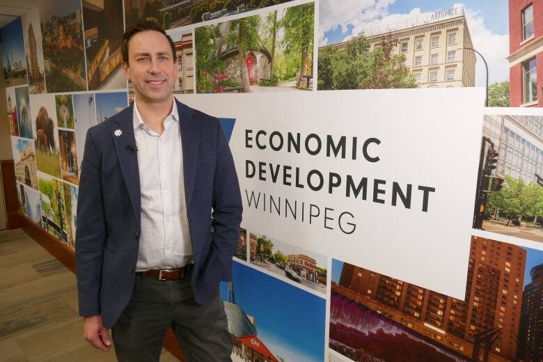 A man in a blue suit and white shirt stands in front of a wall with an assortment of colourful photos of life in Winnipeg.