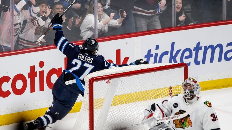 Fans cheer behind the glass behind the Chicago net while a Winnipeg Jets player skates past with his stick raised in celebration.