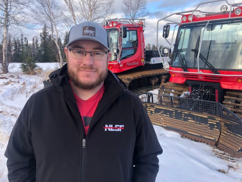 A smiling man wearing a black jacket stands in front of two snow plows.