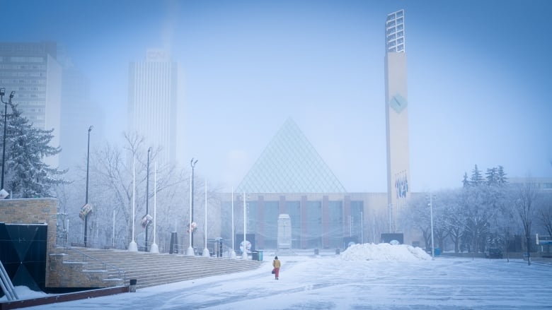 A person walks through an open square covered in snow. The sky is icy blue.