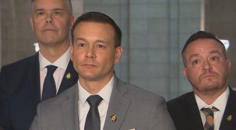 A man in a suit stands at a microphone. Two other men in suits stand behind him.