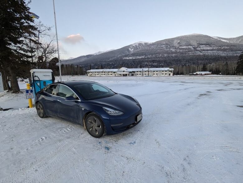 A blue Tesla is plugged in to a charging port in an empty snow covered field. 