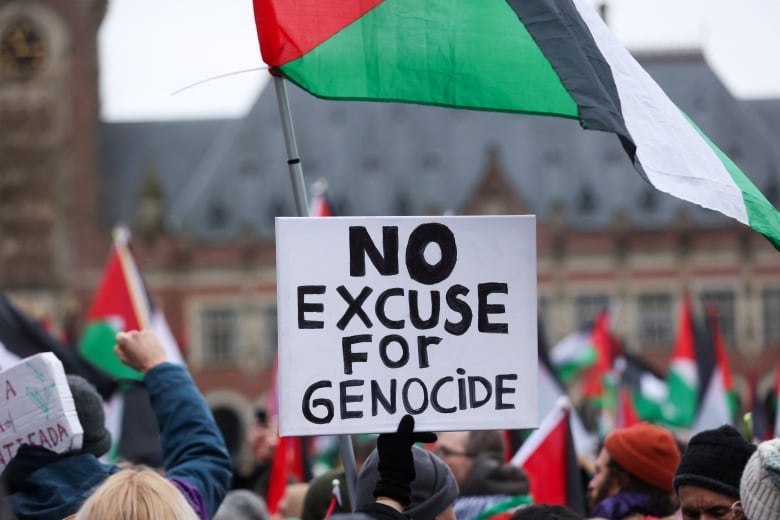 A poster reading 'No excuse for genocide' is held in front of a brick building, with flags waving.