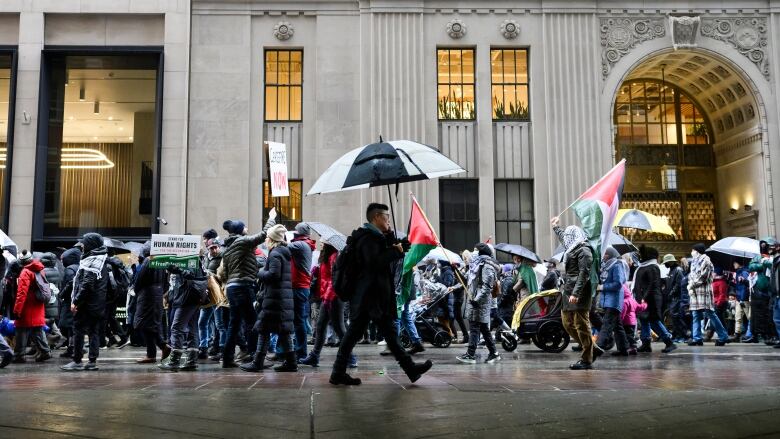 A man carrying an umbrella walks past a group of protesters carrying flags and signs in a city street. 