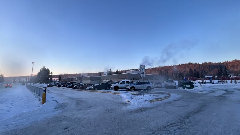 Cars are lined on a parking lot, surrounded by snow and icy roads, and what looks like a small trail of steam is seen above a one-storey building in the background. 