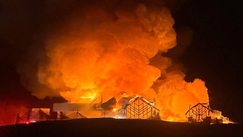 Large plumes of smoke are saturated orange as flames consume a burning golf clubhouse in the night. Emergency lights flash to the left of the building.