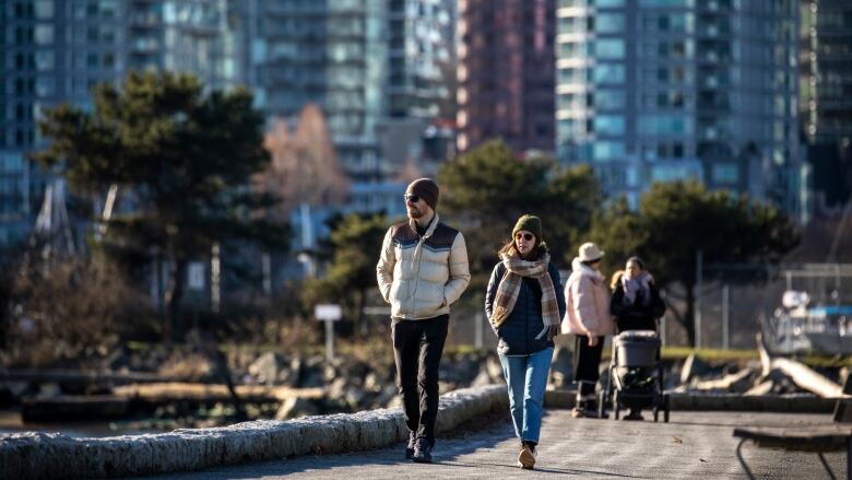 A man in a dark-coloured toque and cream-coloured puffer jacket and a woman in a brown-and-white checkered scarf, dark jacket and green toque are seen walking along a seawall, behind them two other people similarly covered up, standing next to a stroller, and the facades of high-rise buildings in the background.