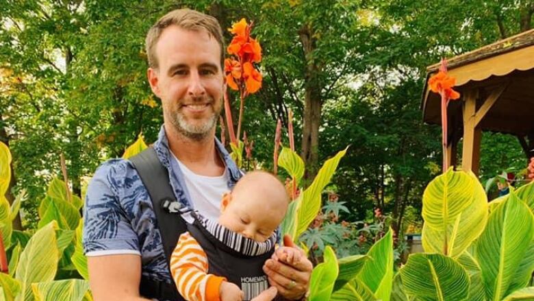 A man smiles at the camera while holding a baby in a baby carrier 