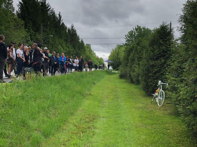 People line up next to a ditch on the side of the road. A white bike rests on the side of the road. 