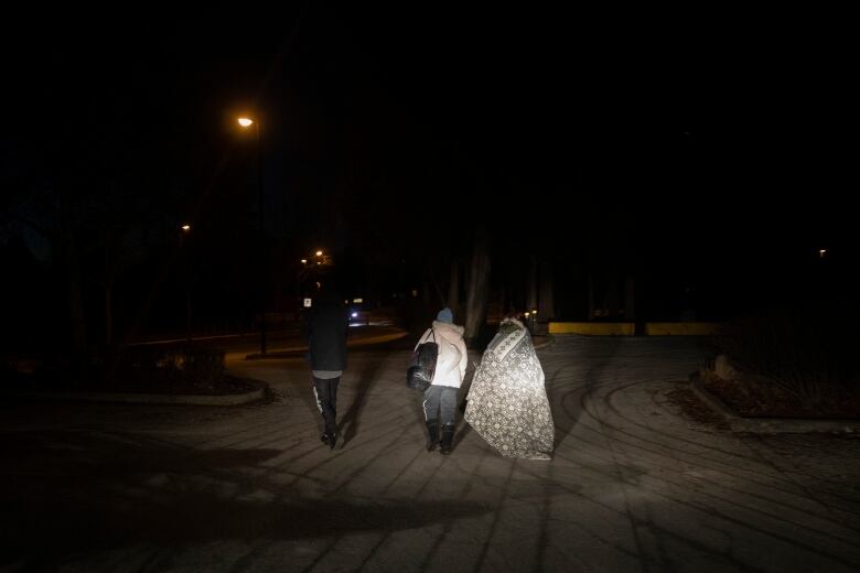 Three people walk side-by-side in the dark facing away from the camera. On the right, the person is draped in a blanket. The person in the middle wears a white coat and carries a handbag and the walker on the left wears all black save faintly visible white writing on the left leg of their pants.