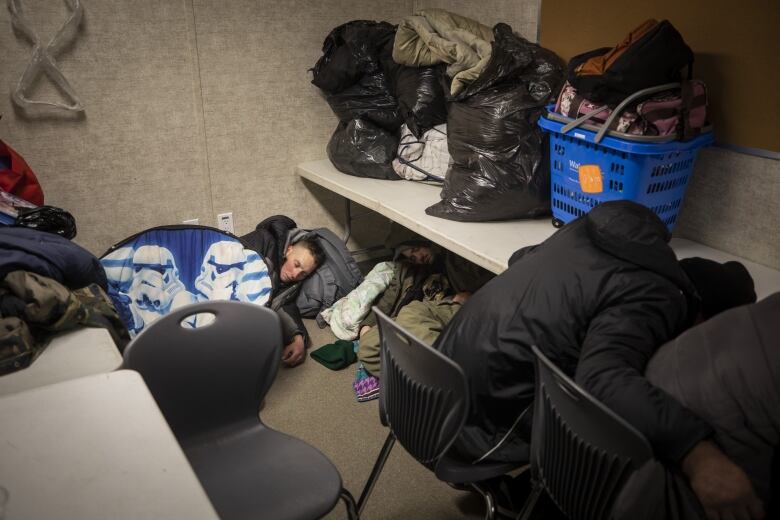 A man sleeps on the floor with his head against a grey backpack. Next to him are men slumped in chairs.