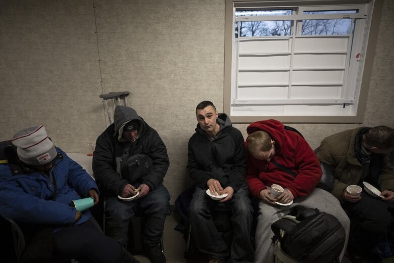 Five men sit in chairs with their backs against a wall. Four are attempting to sleep with their heads slumped over, one white male stares directly at the camera. All of them are holding paper dishes.