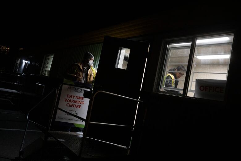 A man stands silhouetted in a trailer doorway that is slightly ajar. He wears a medical mask, toque and safety vest. A young South Asian man is visible inside the window of the trailer also wearing a safety vest. A sign on the exterior of the trailer says Daytime Warming Centre.