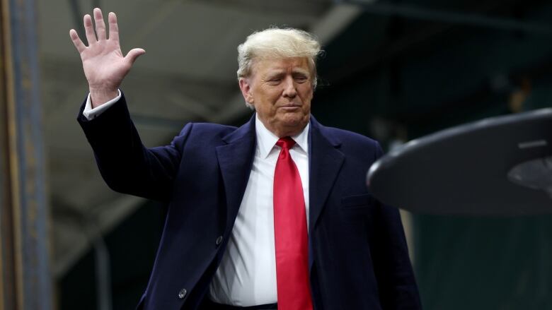 Donald Trump in a dark blue suit and red tie lifts his right hand to wave. 