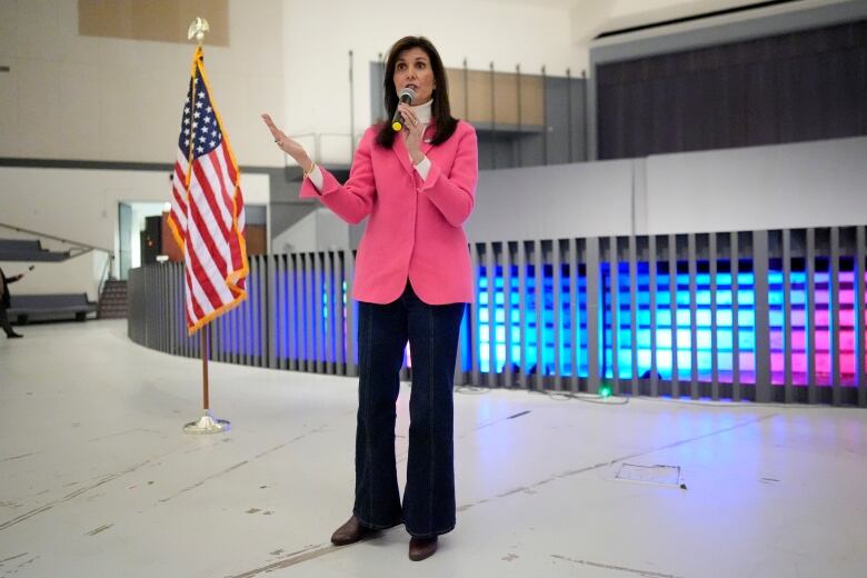 A woman wearing a pink blazer and blue jeans holds a microphone to her mouth while speaking in a room with an American flag behind her. 