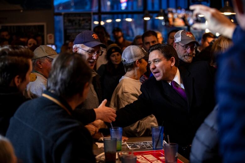 A man in a long, dark coat and purple necktie shakes the hand of another ma over a table in a crowded bar. 