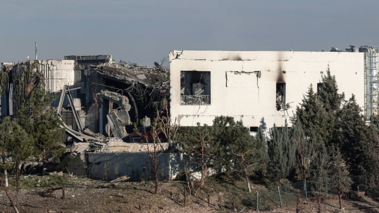 A heavily damaged one-storey structure is shown in a field with collapsed concrete and other debris.