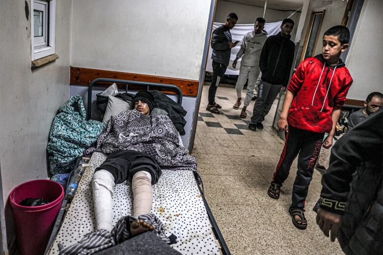 An injured young man lies on a bed in a hospital hallway. Several other people are walking past.