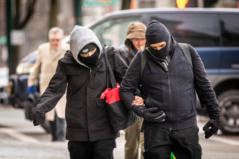 Two people walk with their arms linked. They are both wearing all black as well as balaclavas on their faces as they cross a street with a blue van and other pedestrians in the background.