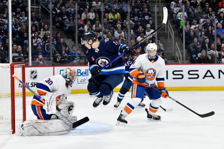 A Winnipeg Jets player in navy blue hops in the air in front the New York Islanders goalie in white.