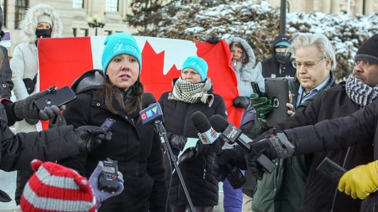 A woman in a black jacket and a blue hat speaks with media. 