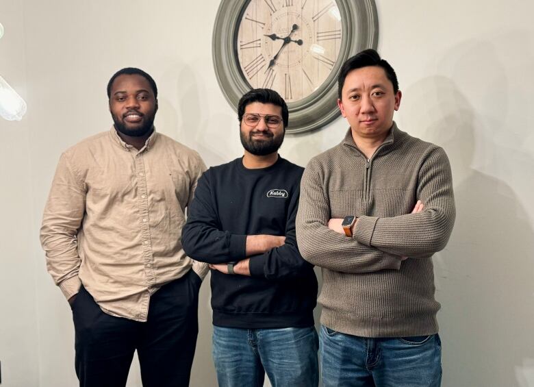 Three men stand next to each other smiling. The man on the far left has his hands in his pockets, while the other two stand with their arms crossed.