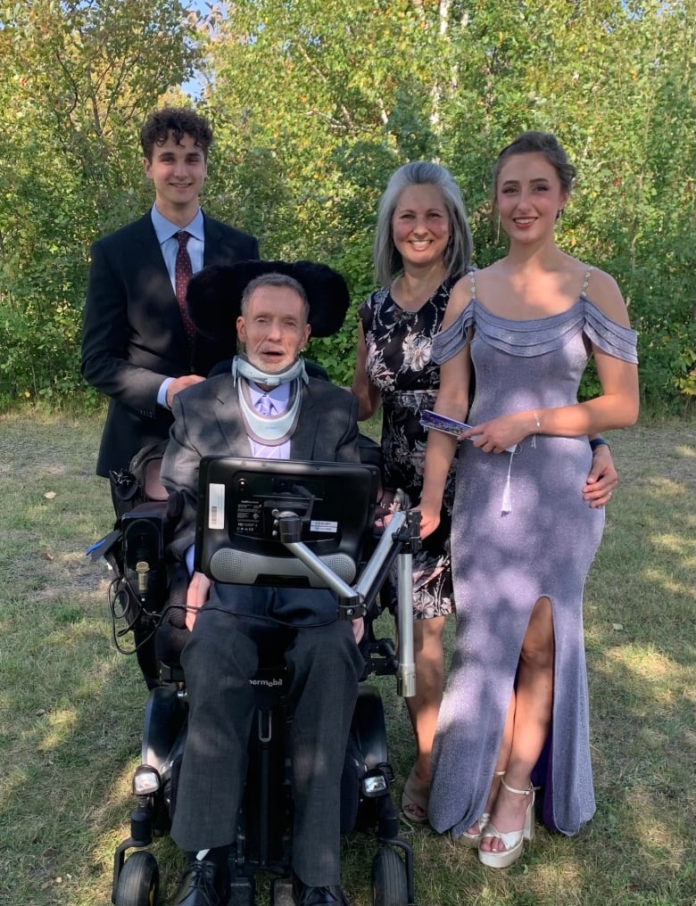 A man in an electric wheelchair is flanked by two young adult children and his wife on a lawn with trees in the background.