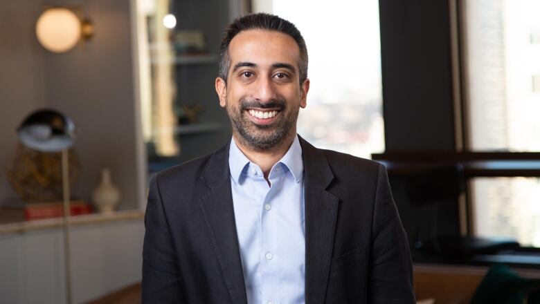 A portrait of a man smiling at the camera. He's dressed in a black blazer and a light blue shirt.