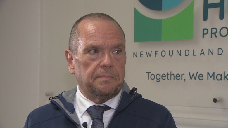 A man with a buzzcut wearing a navy jacket stands in an office space.