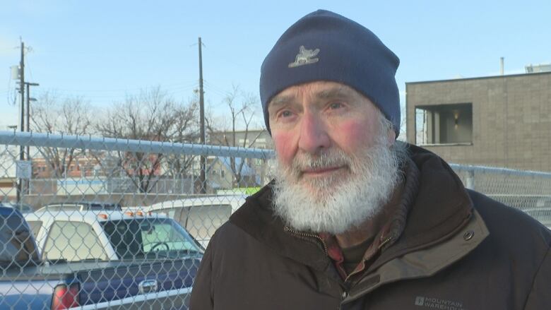 A man with a large white beard and wearing a coat and toque, stands outside in cold weather.