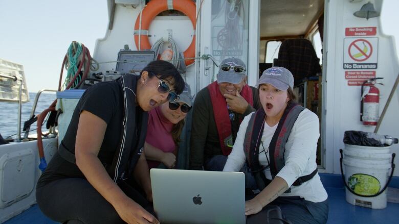 Four people sit on a boat and look at a computer screen with shocked looks on their faces.