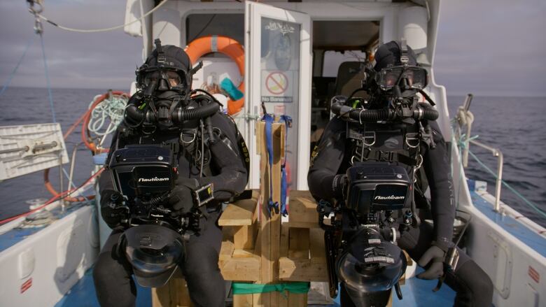 Two men wearing full scuba gear and holding cameras sit patiently on the deck of a fishing boat. 