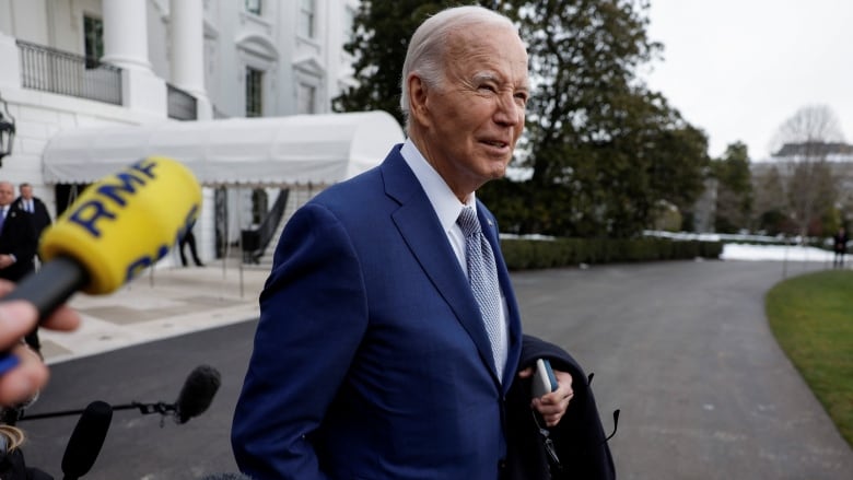 A clean-shaven older man in a suit and tie, holds a coat draped over his arm in front of the White House.