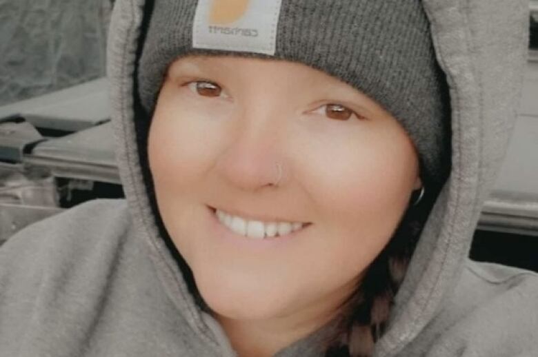 A woman with a smile on her face poses for a picture, wearing a light grey CAT hoodie, and darker grey toque.