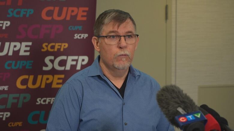 A white man wearing a blue shirt stands in front of a board reading 'SCFP CUPE' and speaks into some microphones.