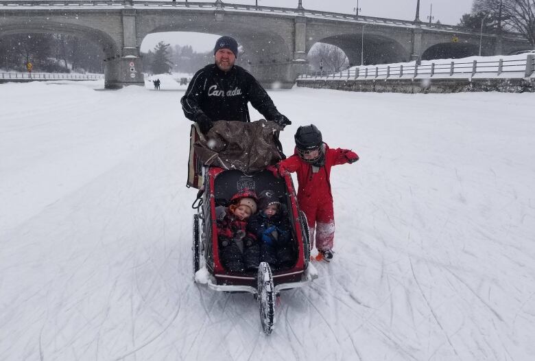 A man wearing a black shirt and grey hat pushes a red stroller along the Rideau Canal Skateway ice with two young children sitting inside. To the right of the stroller is another child wearing a red jumpsuit skating on the ice.