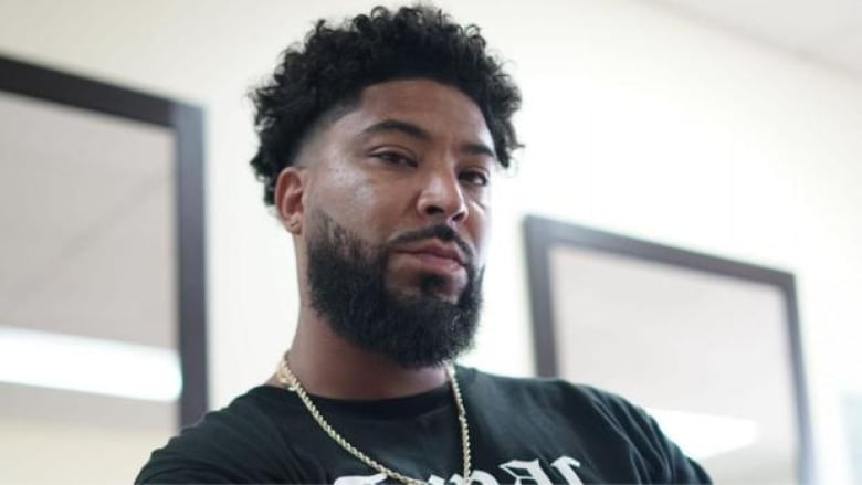 A mid-30s Black man with a beard sitting down wearing a t-shirt with Tupac on it.