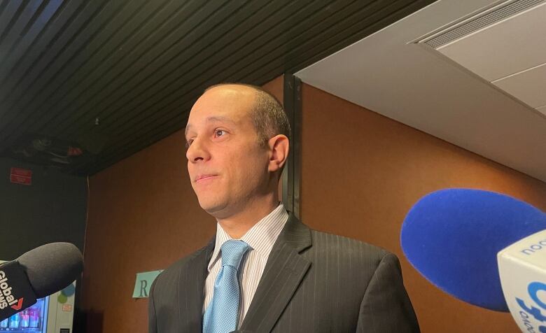 Man seen from three-quarter profile with many media microphones held in front of him.