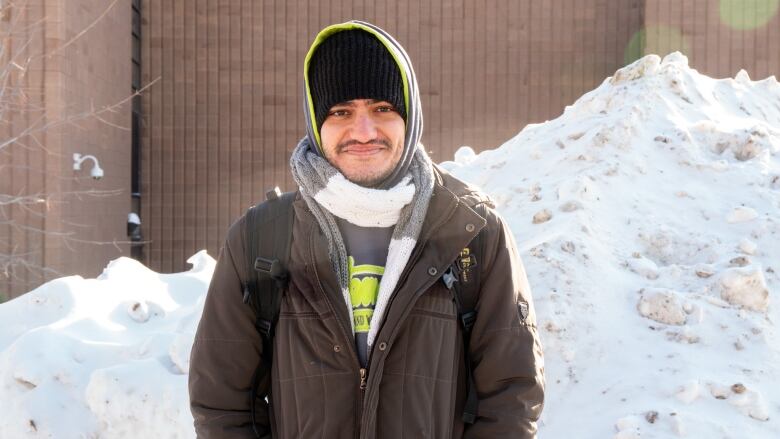 A person wearing a hat and coat with their hood up stands by a large snow bank outside.