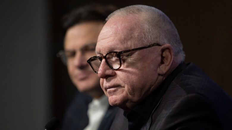 An older white man wearing glasses and a dark suit sits next to a man wearing glasses and a black suit.