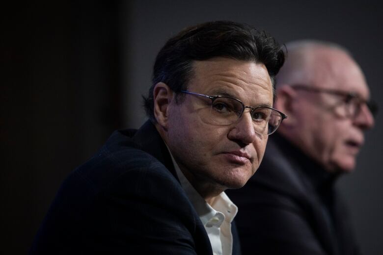 A white man with black hair and wearing glasses and a black suit stares into the camera. An older white man speaks behind him.