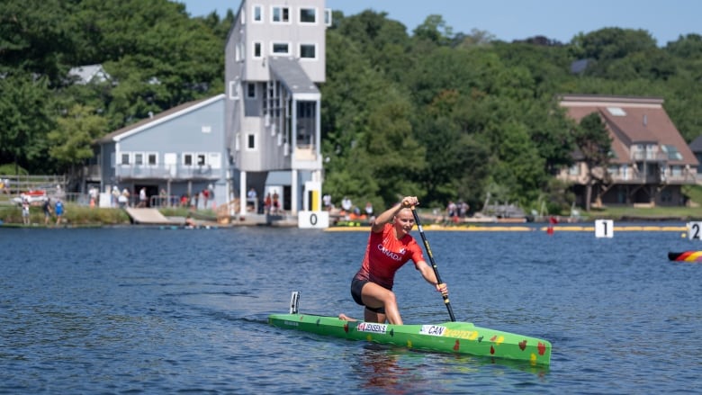 A white woman kneels as she paddles a green kayak across blue water on the lake. The grey two-story judges tower is seen behind her