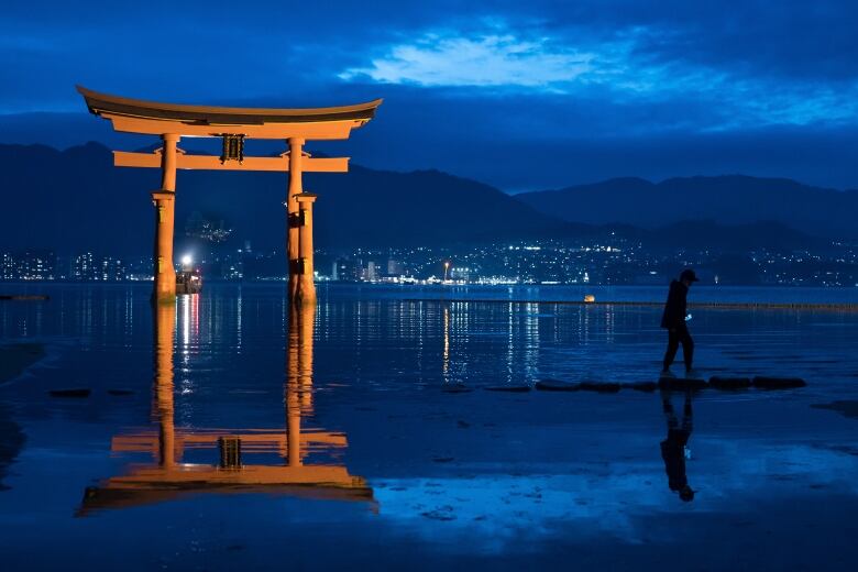 A man walks past a giant East Asian gate located in a water body, with a skyline off in the distance.