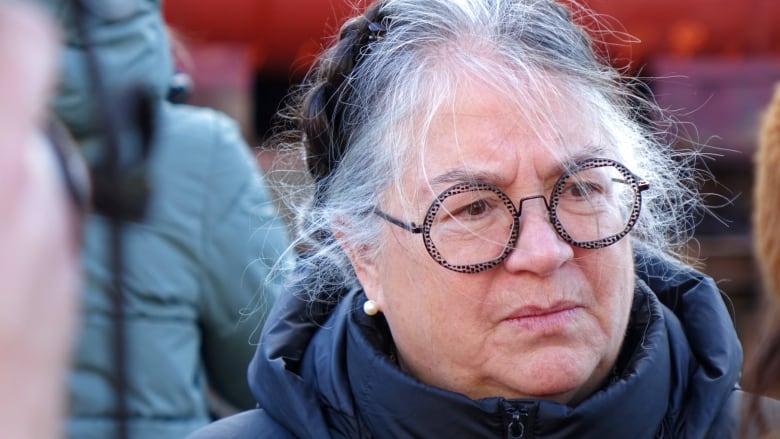 A woman looks concerned outside. She is wearing glasses and her hair is up in a bun.