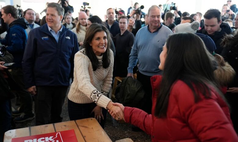 A woman with long brown hair, wearing a white sweater, shakes hands with a woman wearing a red ski jacket, surrounded by a crowd of people.