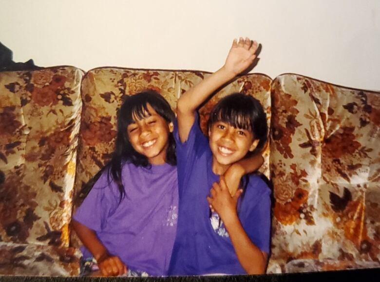 Twin sisters sit closely on a tan couch as children. 